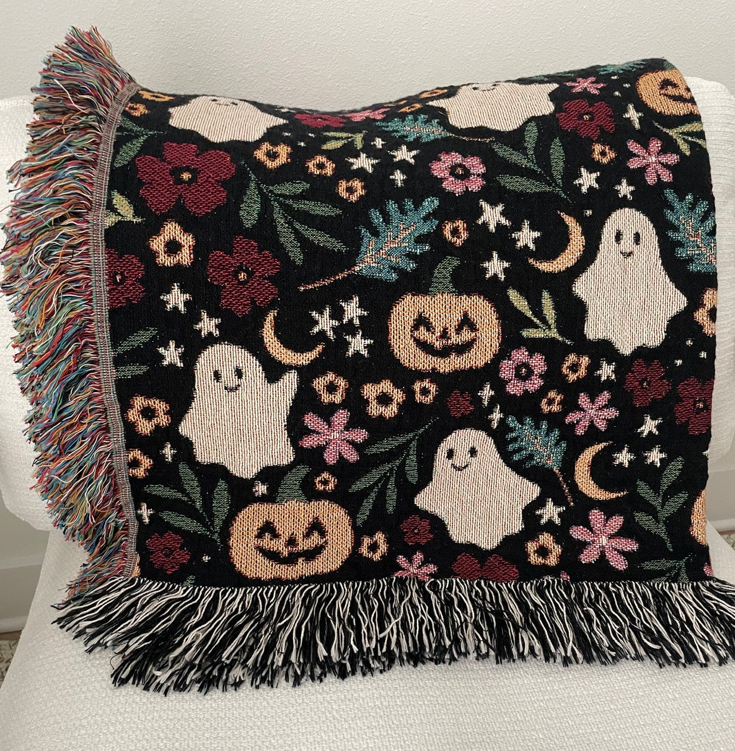 Folded Halloween throw blanket draped over a white chair, showcasing a woven design with smiling ghosts, jack-o’-lanterns, stars, crescent moons, leaves, and florals. Finished with colorful fringe on one side and black-and-white fringe on the bottom.
