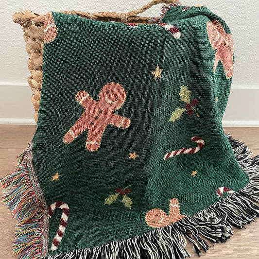 Dark green Christmas woven blanket featuring gingerbread men, candy canes, holly leaves, and multicolored plus black-and-white fringe, draped over a woven basket on a hardwood floor.