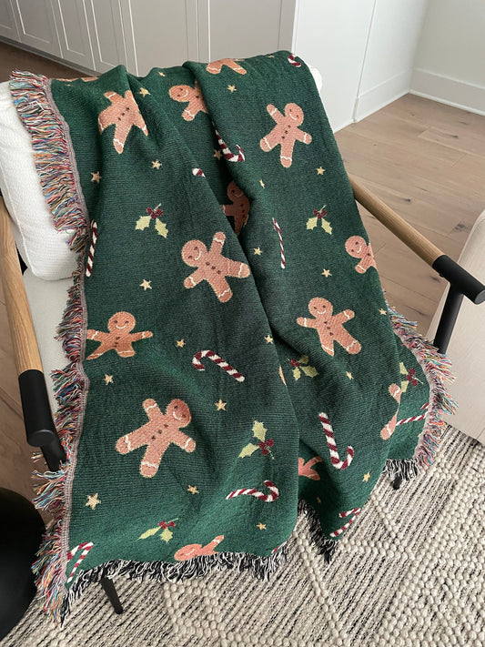 Dark green Christmas woven blanket featuring gingerbread men, candy canes, holly leaves, and multicolored plus black-and-white fringe, draped over a white armchair.