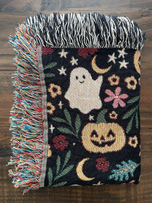 Folded Halloween woven throw blanket on a wooden surface, featuring a whimsical design with ghosts, jack-o’-lanterns, crescent moons, stars, and florals. Colorful fringe on one side and black-and-white fringe on the other.