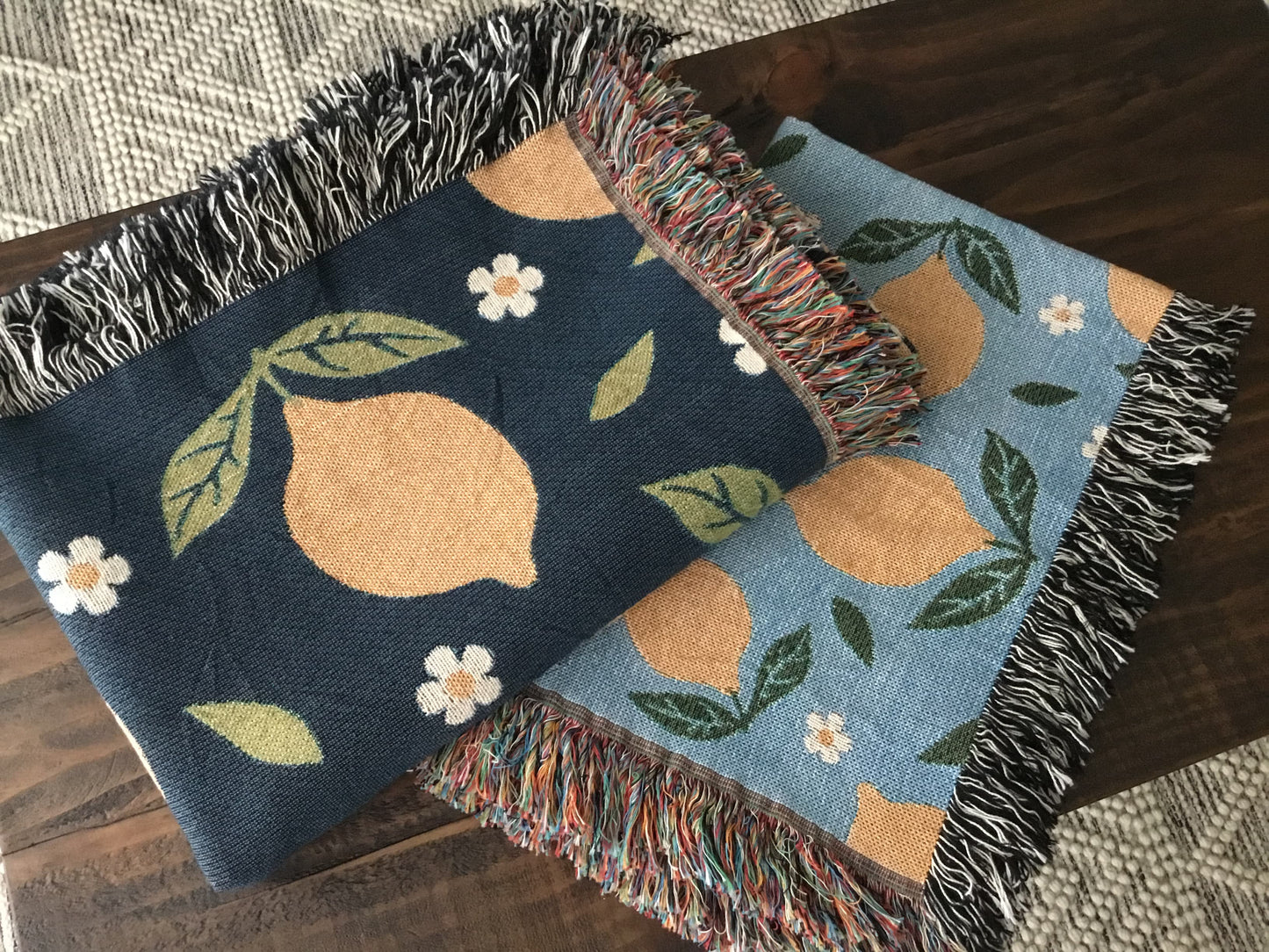 Two lemon-themed woven throw blankets with daisy patterns—one in navy and one in light blue—featuring fringe edges, folded on a wooden surface.