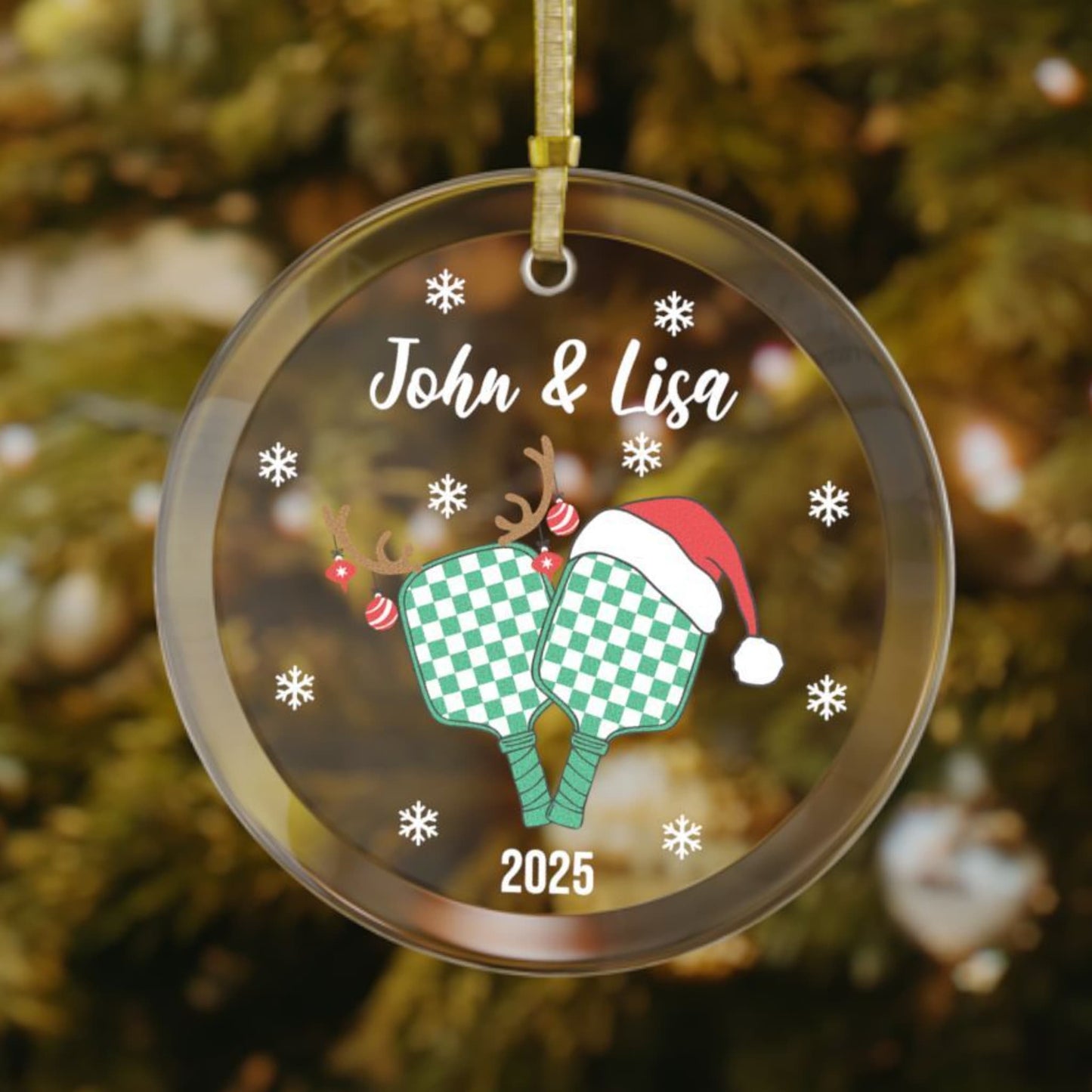 Personalized pickleball couples or teammate Christmas ornament featuring two green checkered paddles, one with antlers and ornaments, the other with a Santa hat, snowflakes surrounding the design, with custom names on top and the year at the bottom.