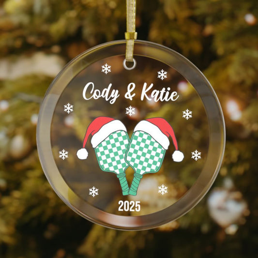 Personalized pickleball couples or teammate Christmas ornament featuring two green checkered paddles wearing Santa hats, snowflakes surrounding the design, with custom names on top and the year at the bottom.
