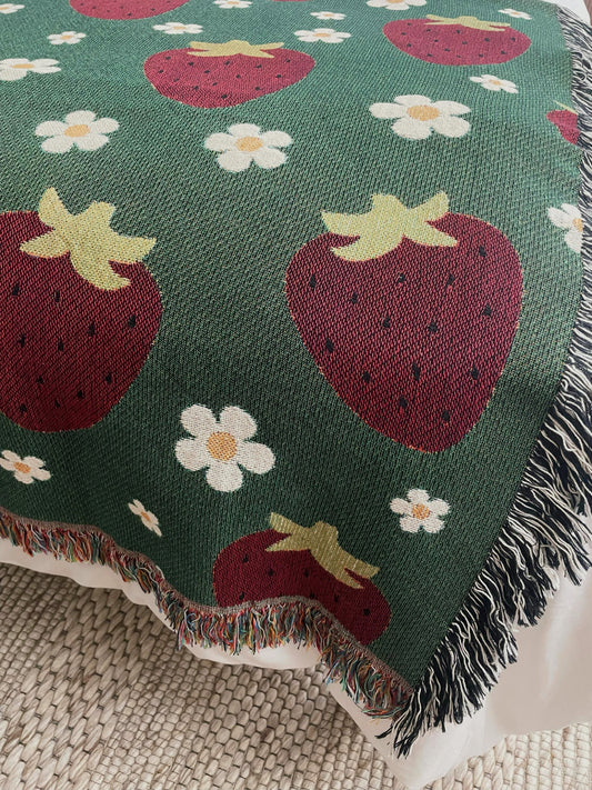 Green woven throw blanket with red strawberries and white daisies, featuring multicolored and black-and-white fringe, styled on a bed for cottagecore or farmhouse decor.