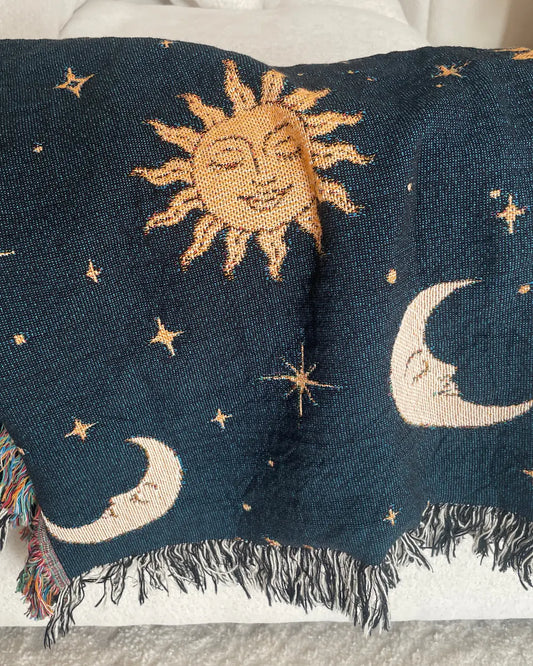 Celestial woven throw blanket with golden sun faces, cream crescent moons and scattered stars on a dark navy background, draped over a white sofa with colorful and black and white fringe edges.