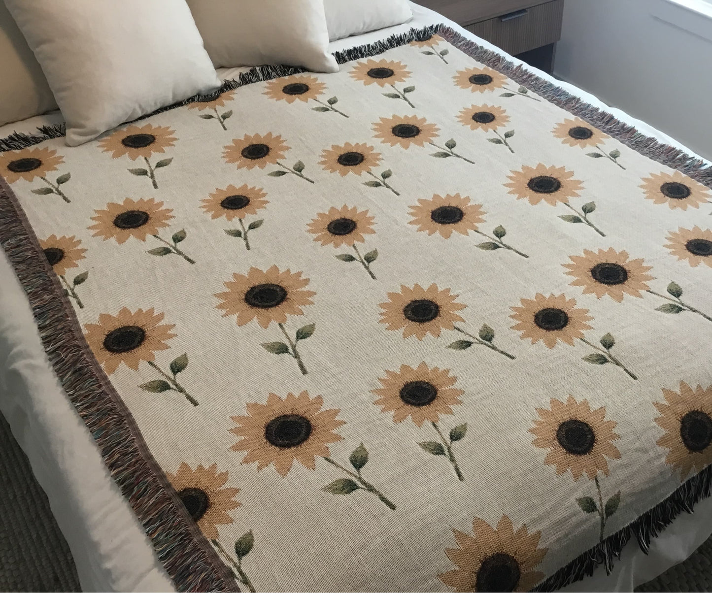 Beige woven sunflower throw blanket laid flat on a neatly made bed, featuring a repeating sunflower pattern with multicolored and black-and-white fringe edges, styled with beige pillows for a cozy, boho farmhouse-inspired look.