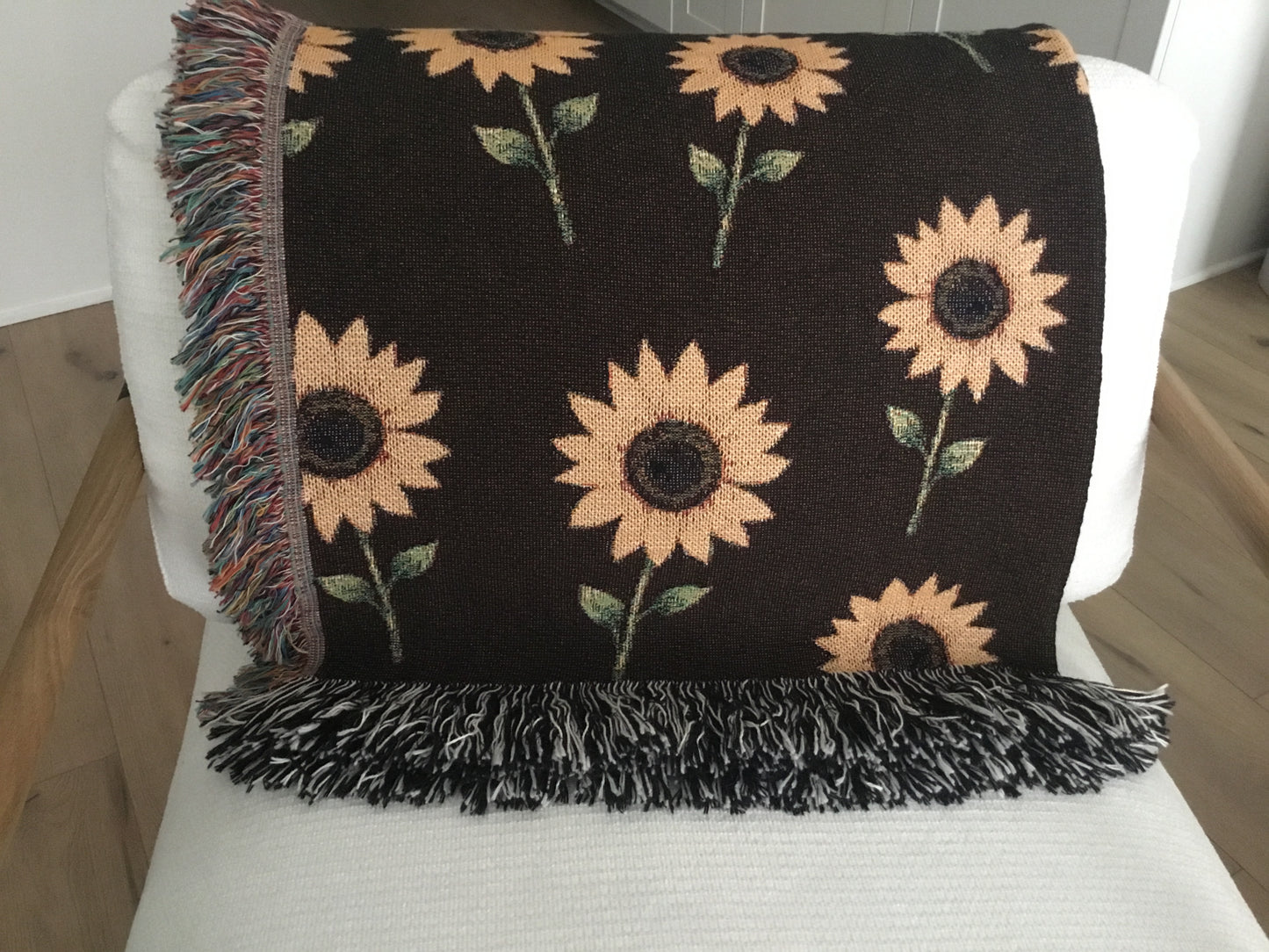 Dark brown woven sunflower throw blanket featuring a yellow sunflower pattern with multicolored fringe on the sides and black-and-white fringe on the top and bottom, neatly draped over the back of a white armchair.