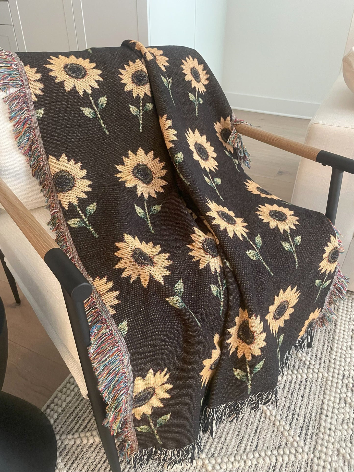 Cozy woven sunflower throw blanket draped over a modern wood-accent armchair on a textured cream rug, featuring a yellow sunflower pattern on a dark brown background with colorful fringe on one side and multicolored fringe on the other.