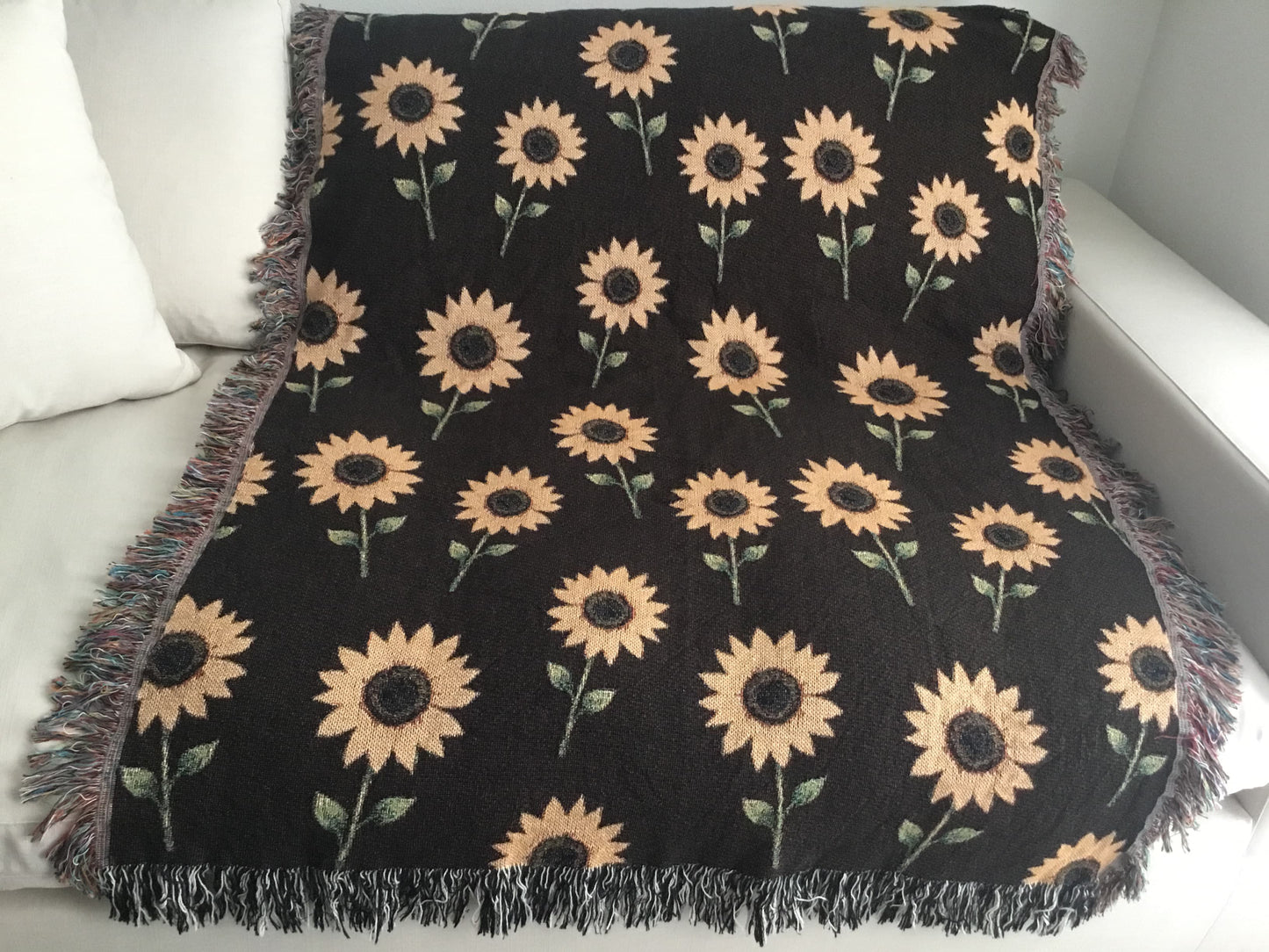 Dark brown woven sunflower throw blanket laid flat over a white couch, showcasing a repeating yellow sunflower pattern and multicolored and black and white fringe edges.