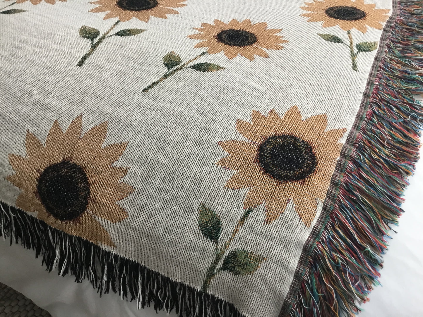 Close-up of a beige woven sunflower throw blanket featuring a repeating pattern of sunflower plants with dark brown centers, green leaves, and stems, finished with multicolored fringe on the sides and black-and-white fringe along the top and bottom edges.