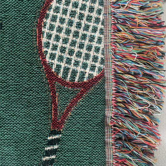 Close-up of a green woven tennis racket throw blanket showing detailed stitching and multicolor fringe.