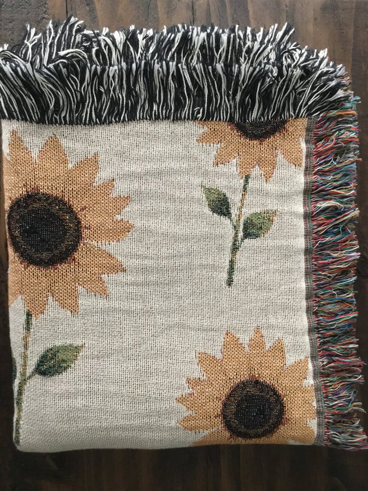 Folded beige woven sunflower throw blanket displayed on a dark wood surface, featuring a pattern of sunflower plants with dark brown centers, green leaves, and multicolored fringe on the side and black-and-white fringe along the top edge.