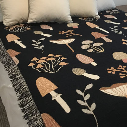 Black woven throw blanket with mushroom and foliage motifs, styled on a bed with cream pillows — cozy cottagecore or woodland decor.