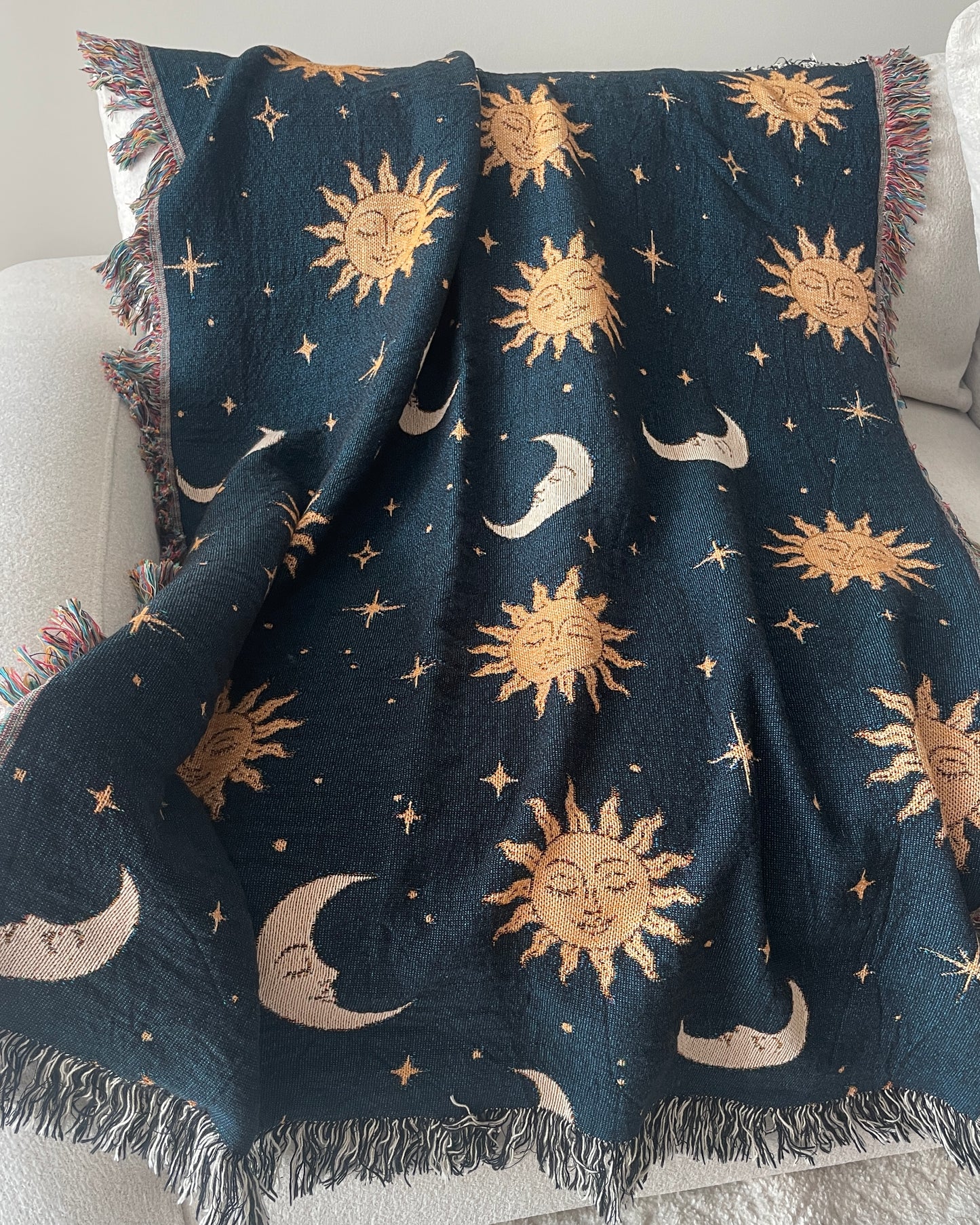 Celestial woven throw blanket with golden sun faces, crescent moons and scattered stars on a dark navy blue background, draped over a white sofa with fringed edges.