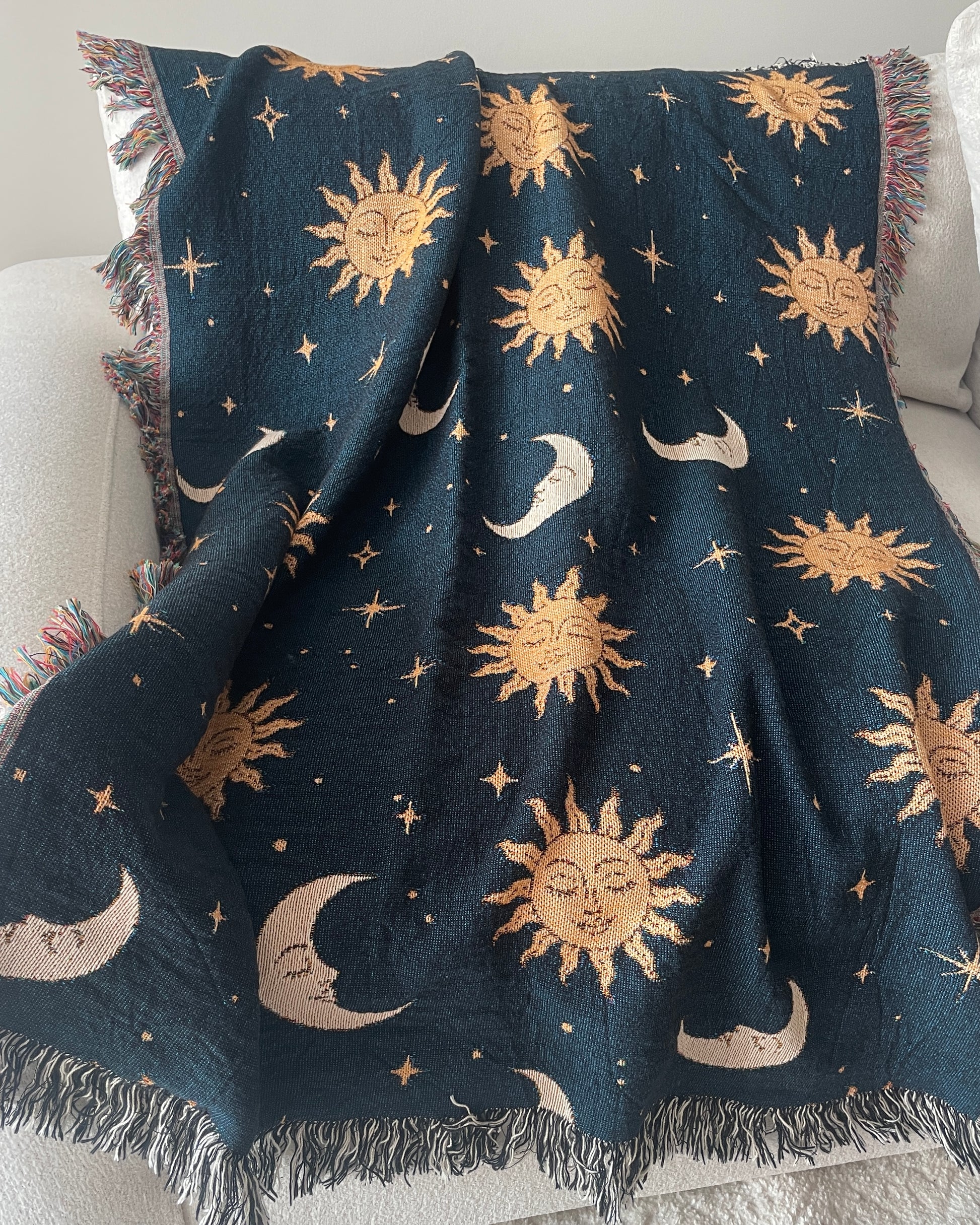 Celestial woven throw blanket with golden sun faces, crescent moons and scattered stars on a dark navy blue background, draped over a white sofa with fringed edges.