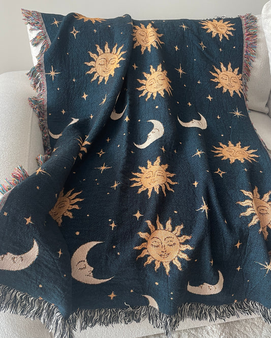 Celestial woven throw blanket with golden sun faces, crescent moons and scattered stars on a dark navy blue background, draped over a white sofa with fringed edges.
