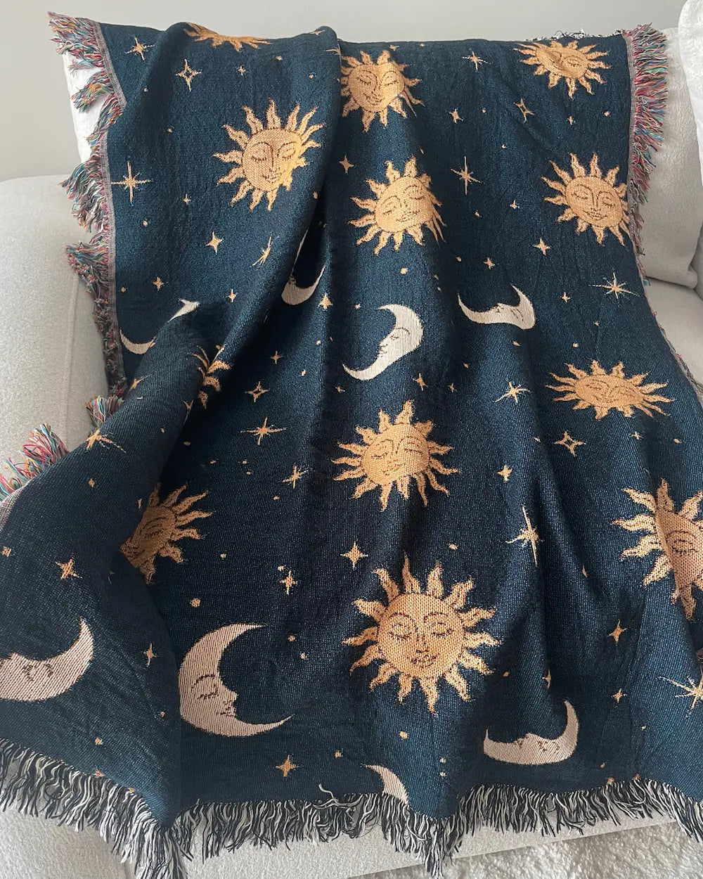 Celestial woven throw blanket with golden sun faces, crescent moons and scattered stars on a dark navy blue background, draped over a white sofa with fringed edges.