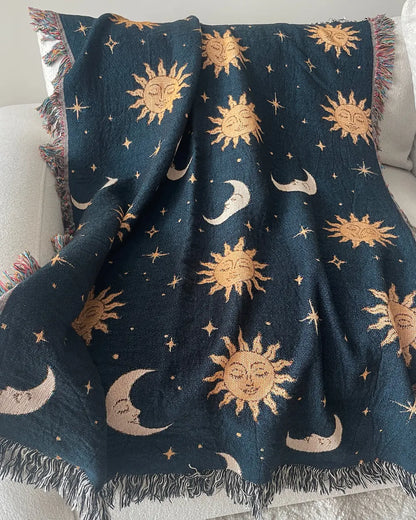 Celestial woven throw blanket with golden sun faces, crescent moons and scattered stars on a dark navy blue background, draped over a white sofa with fringed edges.