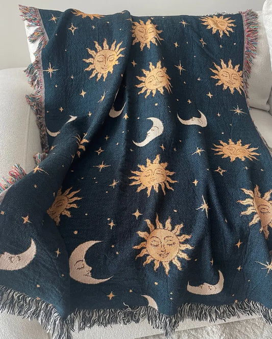 Celestial woven throw blanket with golden sun faces, crescent moons and scattered stars on a dark navy blue background, draped over a white sofa with fringed edges.