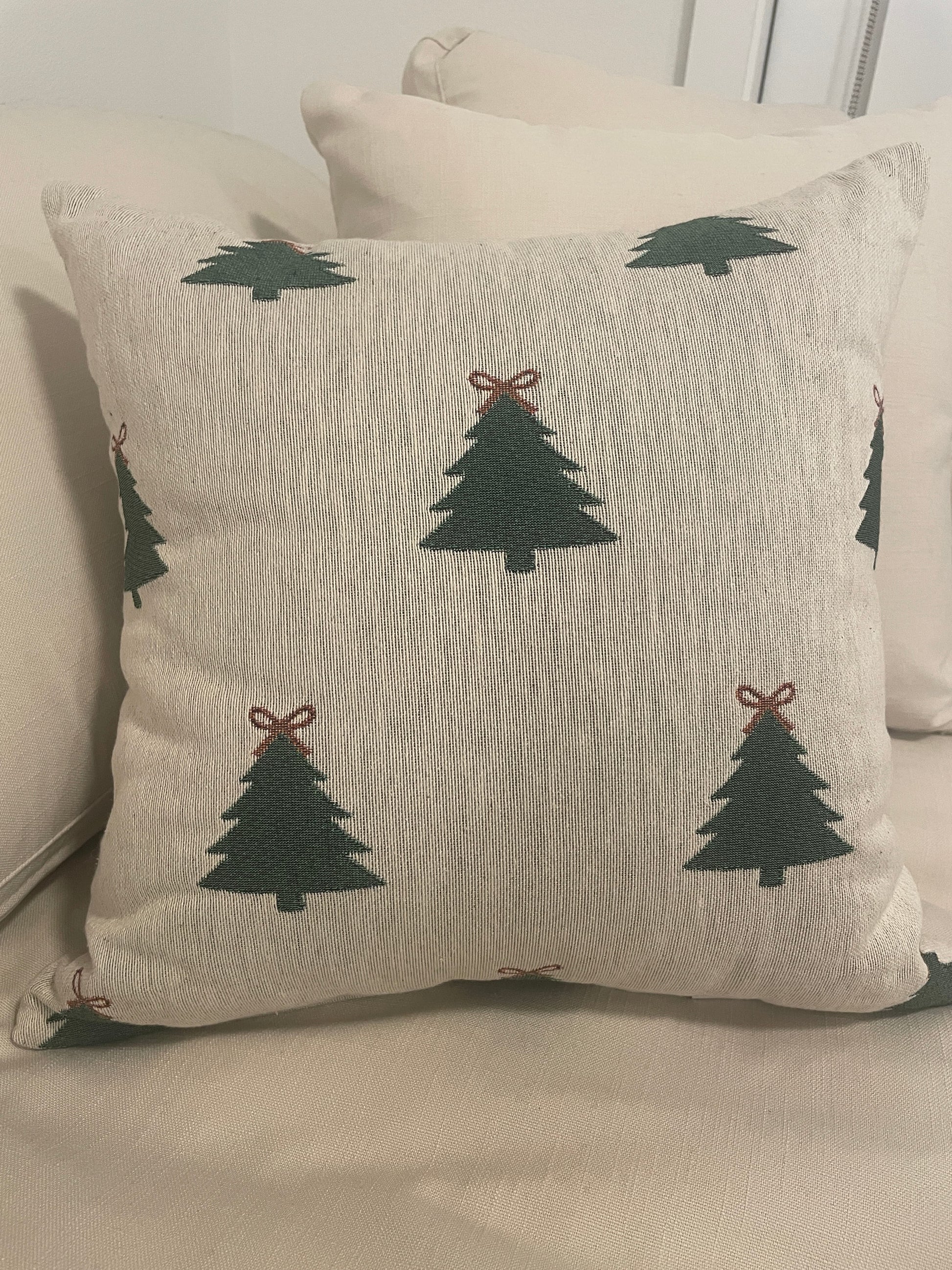 Beige woven Christmas throw pillow with green Christmas tree pattern and red bow accents, styled on a sofa for cozy holiday décor.