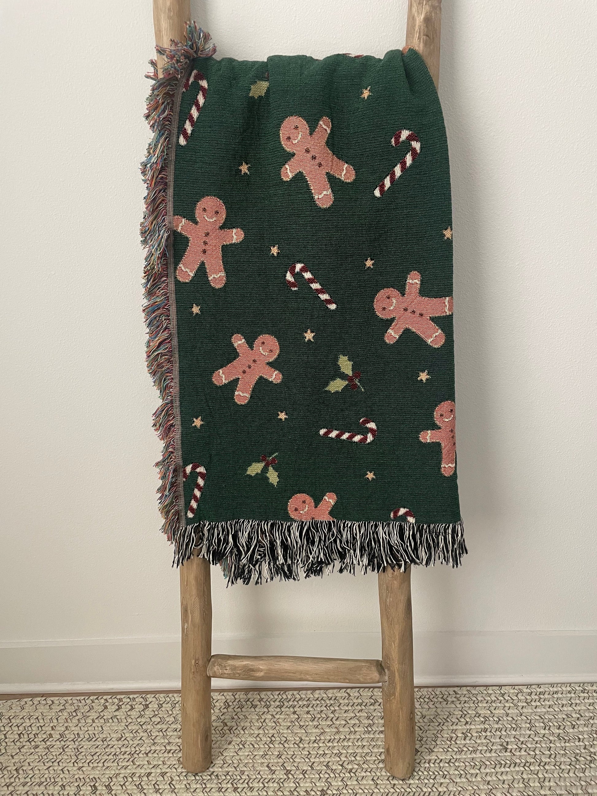 Dark green Christmas blanket featuring gingerbread men, candy canes, and holly, styled on a wooden ladder.