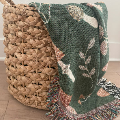 Green woven mushroom throw blanket with colorful fringe draped over a natural wicker basket — cozy cottagecore or woodland home decor accent.