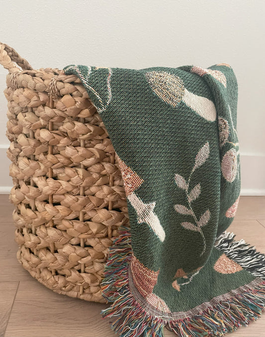 Green woven mushroom throw blanket with colorful fringe draped over a natural wicker basket — cozy cottagecore or woodland home decor accent.