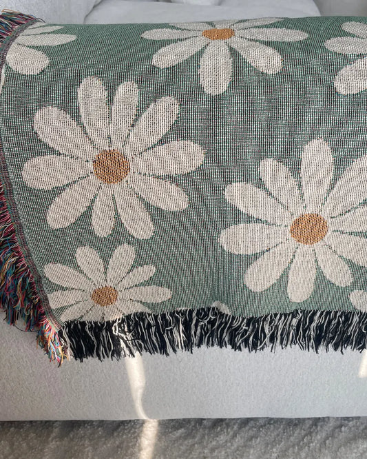 Sage green daisy woven throw blanket draped over a white sofa with cream flowers and multicolor and black-and-white fringe.