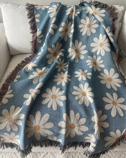 Dusty blue daisy woven throw blanket with oversized daisy flowers and colorful fringe, draped over a white sofa