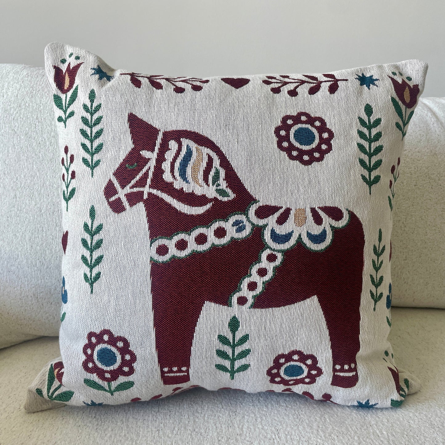 A square throw pillow with a traditional Swedish Dala horse design, featuring a dark red horse and floral motifs on a cream background, styled on a light beige sofa.