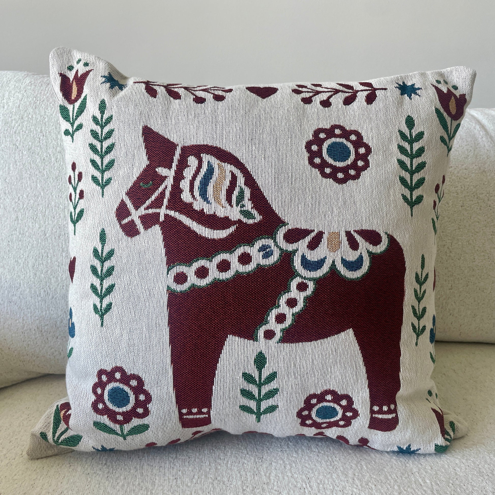 A square throw pillow with a traditional Swedish Dala horse design, featuring a dark red horse and floral motifs on a cream background, styled on a light beige sofa.