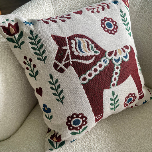 A 17x18 throw pillow with a traditional Swedish Dala horse design, resting on a textured cream-colored armchair.