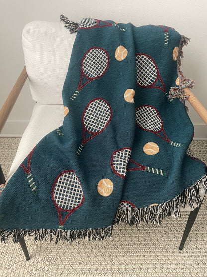Dark teal tennis racket woven throw blanket with fringe draped over a chair.