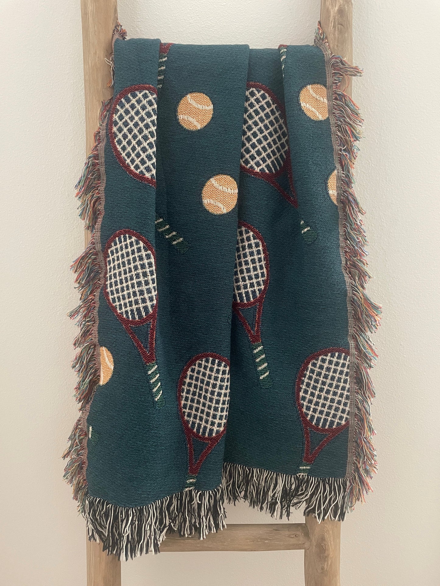 Dark teal tennis racket woven throw blanket with fringe displayed on a wooden ladder.