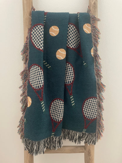 Dark teal tennis racket woven throw blanket with fringe displayed on a wooden ladder.