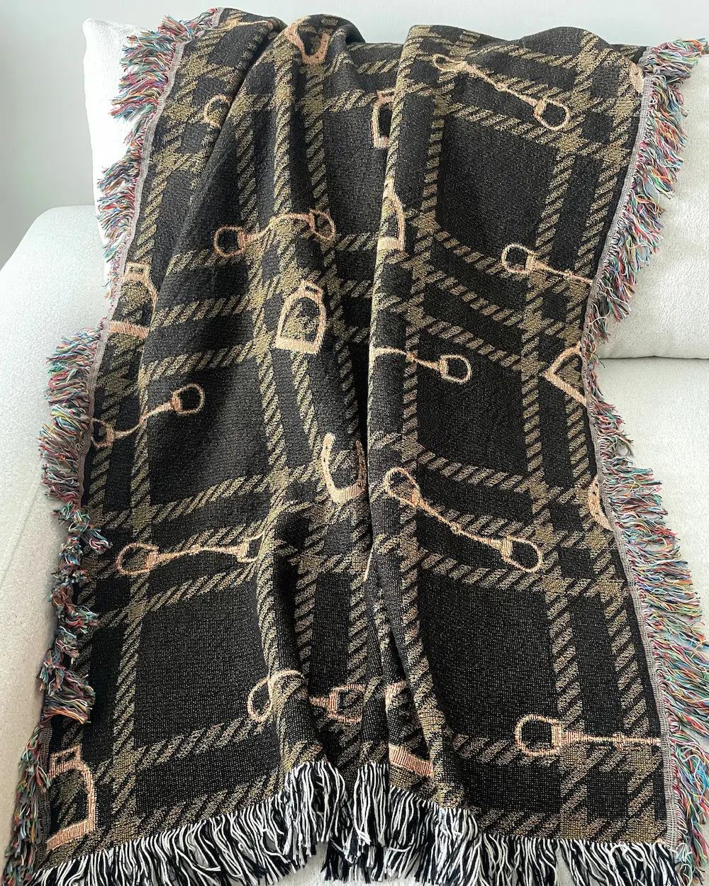 Equestrian woven throw blanket with a dark brown plaid pattern, horse bits and stirrups, and black-and-white and multicolored fringe, draped over a white sofa.