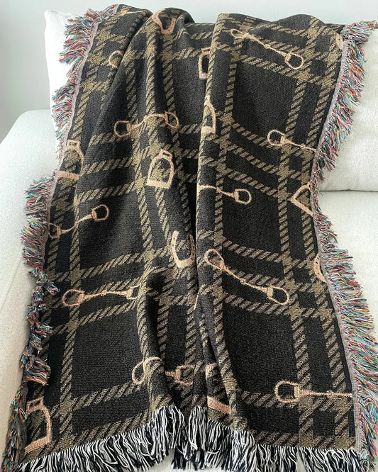 Equestrian woven throw blanket with a dark brown plaid pattern, horse bits and stirrups, and black-and-white and multicolored fringe, draped over a white sofa.
