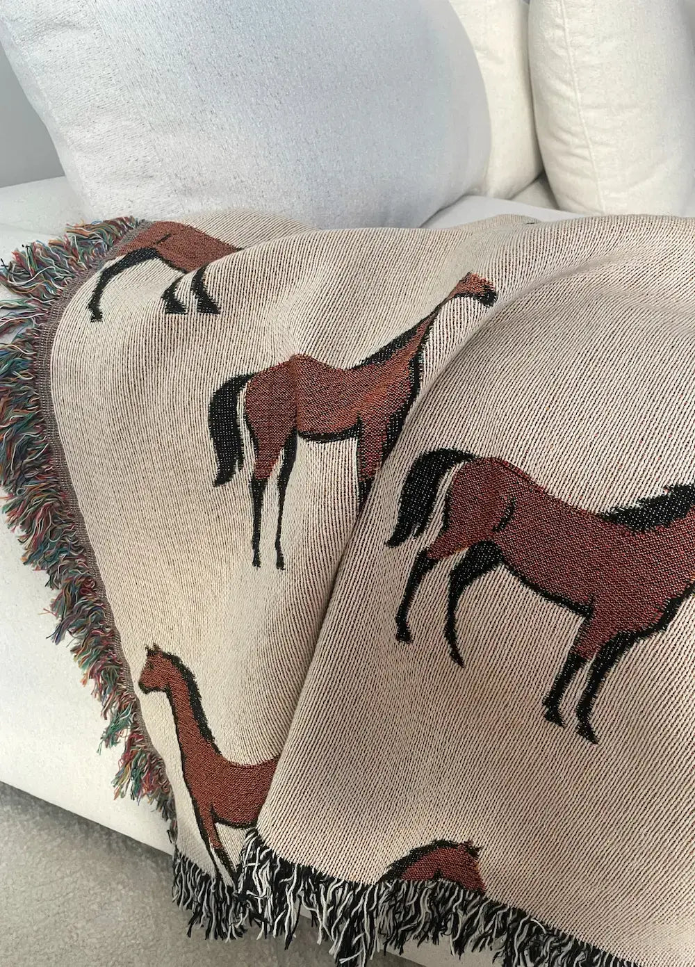 Horse motif woven blanket draped over a neutral sofa, featuring a repeating equestrian pattern in earthy neutrals with fringe edges, jacquard woven in 100% cotton.