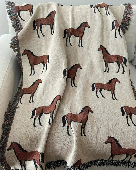 Horse woven throw blanket in cream with an all-over brown horse pattern, black-and-white fringe on one side and multicolored fringe on the other, draped over a light-colored sofa.
