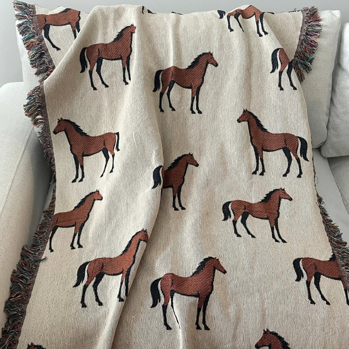 Horse motif woven blanket made from 100% cotton jacquard, featuring repeating chestnut horses on a neutral background with fringe edges, styled on a sofa for classic equestrian home decor.