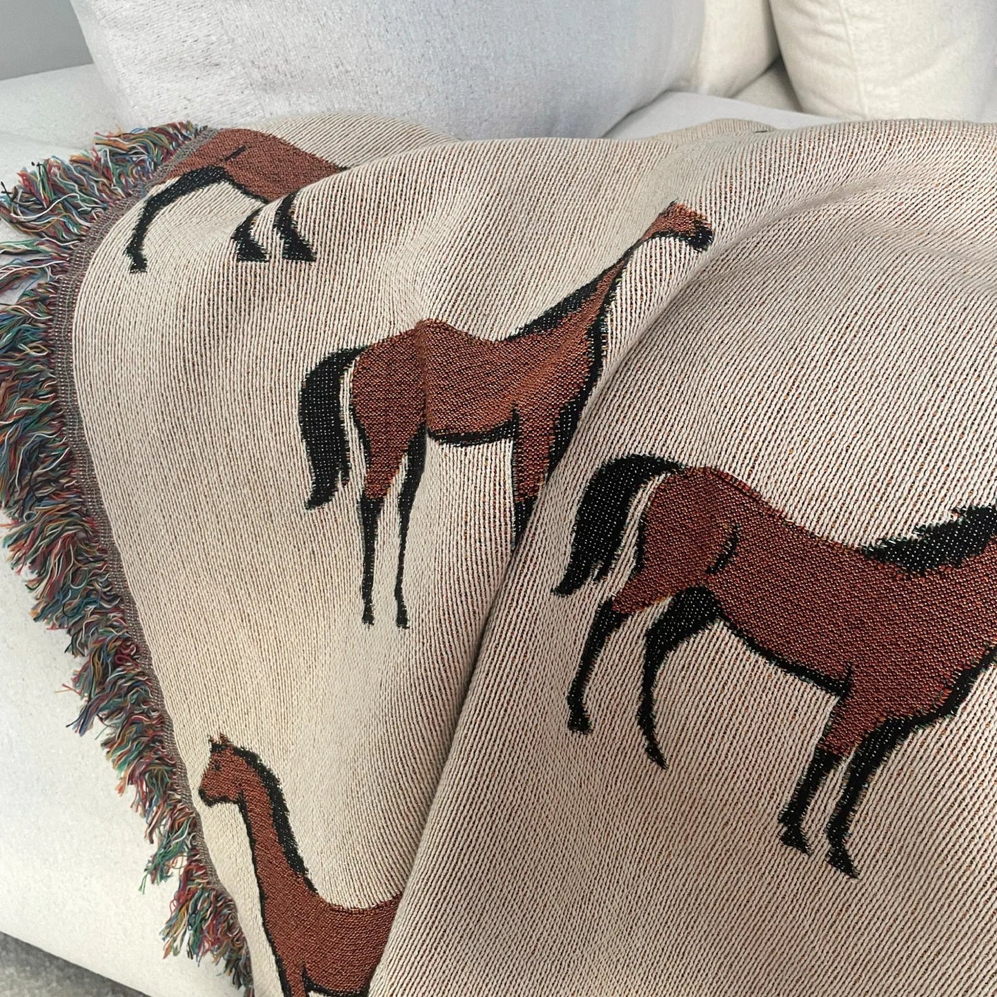 Horse motif woven blanket draped over a neutral sofa, featuring a repeating equestrian pattern in earthy neutrals with fringe edges, jacquard woven in 100% cotton.
