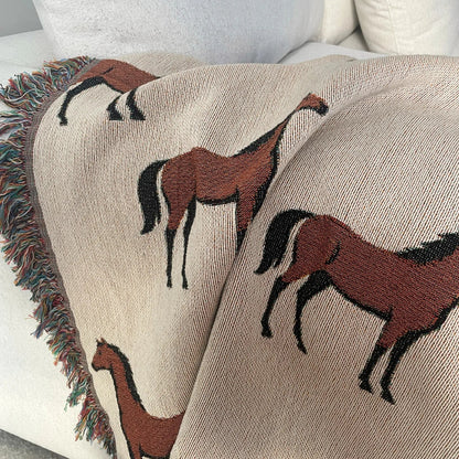 Horse motif woven blanket draped over a neutral sofa, featuring a repeating equestrian pattern in earthy neutrals with fringe edges, jacquard woven in 100% cotton.