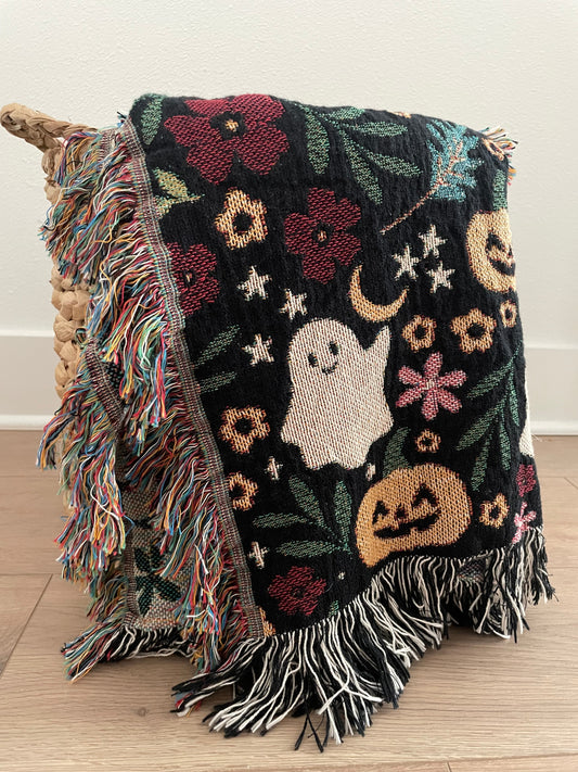 Woven Halloween throw blanket draped over a basket, featuring cute ghosts, jack-o'-lanterns, stars, moons, and floral motifs on a black background with fringe edges.