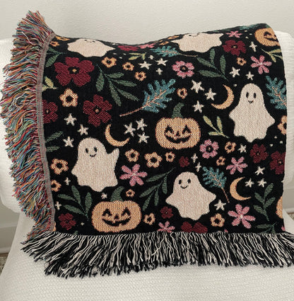 Folded Halloween throw blanket draped over a white chair, showcasing a woven design with smiling ghosts, jack-o’-lanterns, stars, crescent moons, leaves, and florals. Finished with colorful fringe on one side and black-and-white fringe on the bottom.
