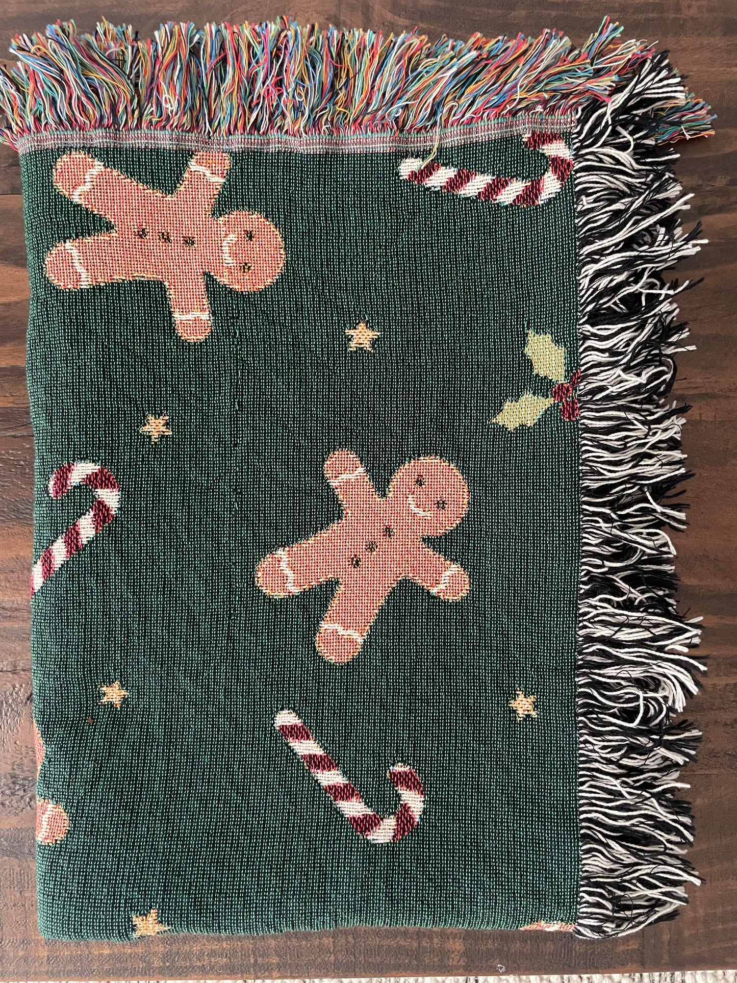Dark green Christmas woven blanket featuring gingerbread men, candy canes, holly leaves, and multicolored plus black-and-white fringe, folded neatly on a dark wood surface.