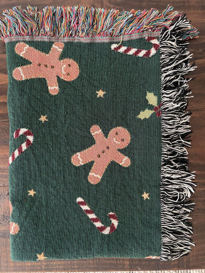 Dark green Christmas woven blanket featuring gingerbread men, candy canes, holly leaves, and multicolored plus black-and-white fringe, folded neatly on a dark wood surface.