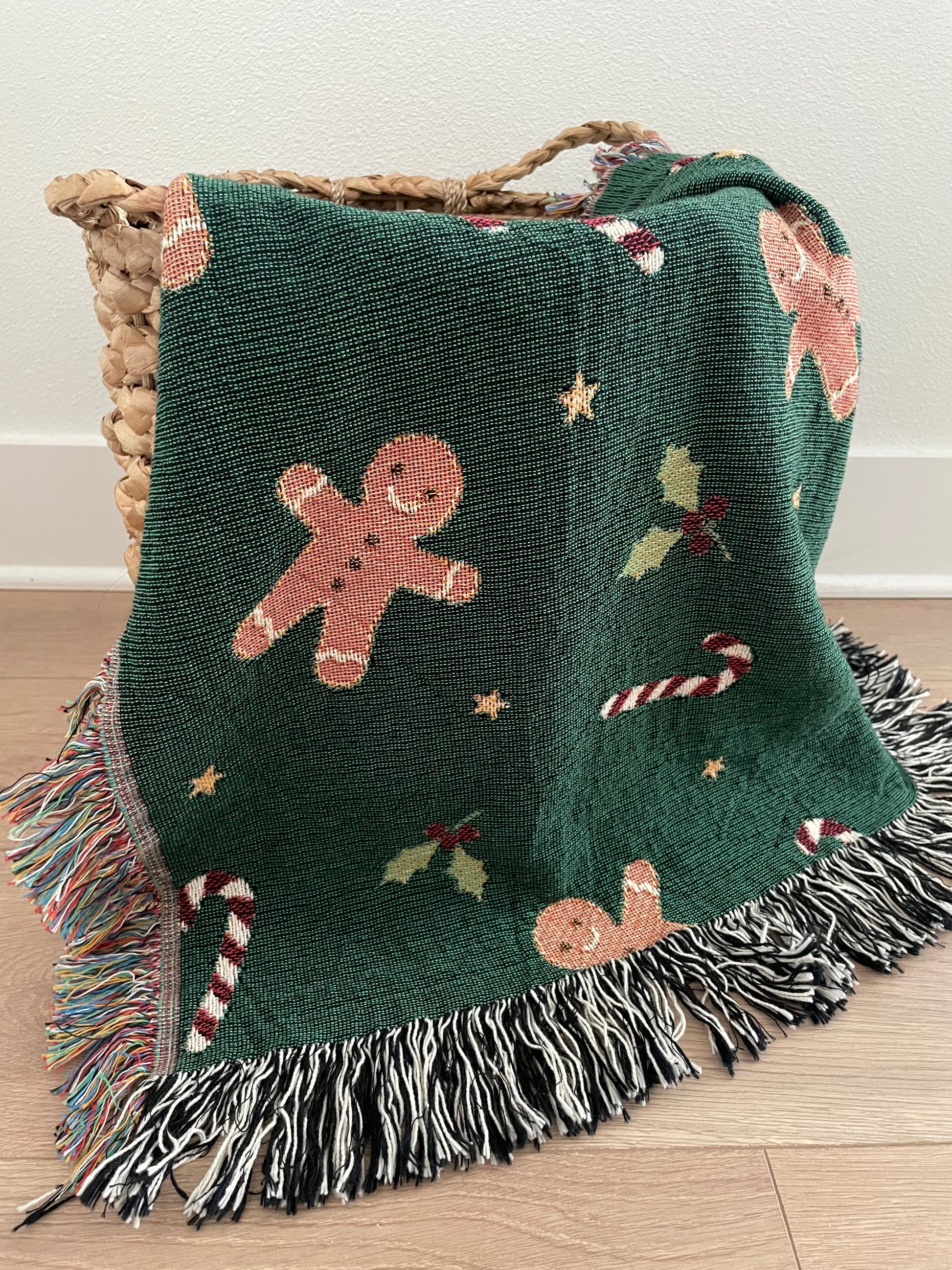 Dark green Christmas woven blanket featuring gingerbread men, candy canes, holly leaves, and multicolored plus black-and-white fringe, draped over a woven basket on a hardwood floor.