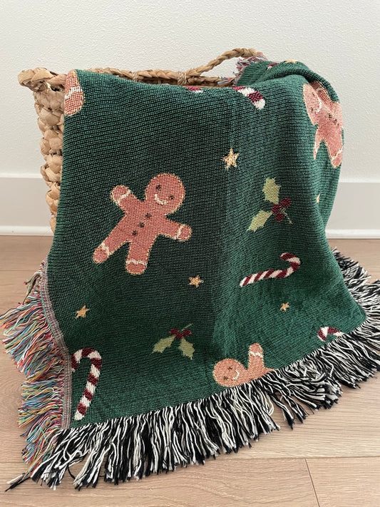 Dark green Christmas woven blanket featuring gingerbread men, candy canes, holly leaves, and multicolored plus black-and-white fringe, draped over a woven basket on a hardwood floor.
