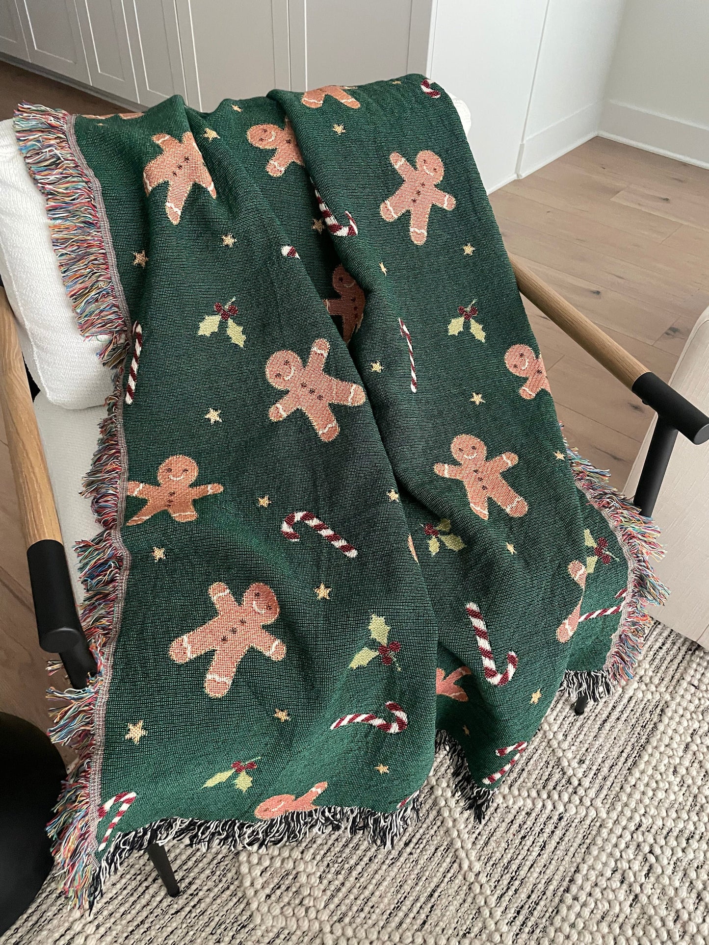 Dark green Christmas woven blanket featuring gingerbread men, candy canes, holly leaves, and multicolored plus black-and-white fringe, draped over a white armchair.