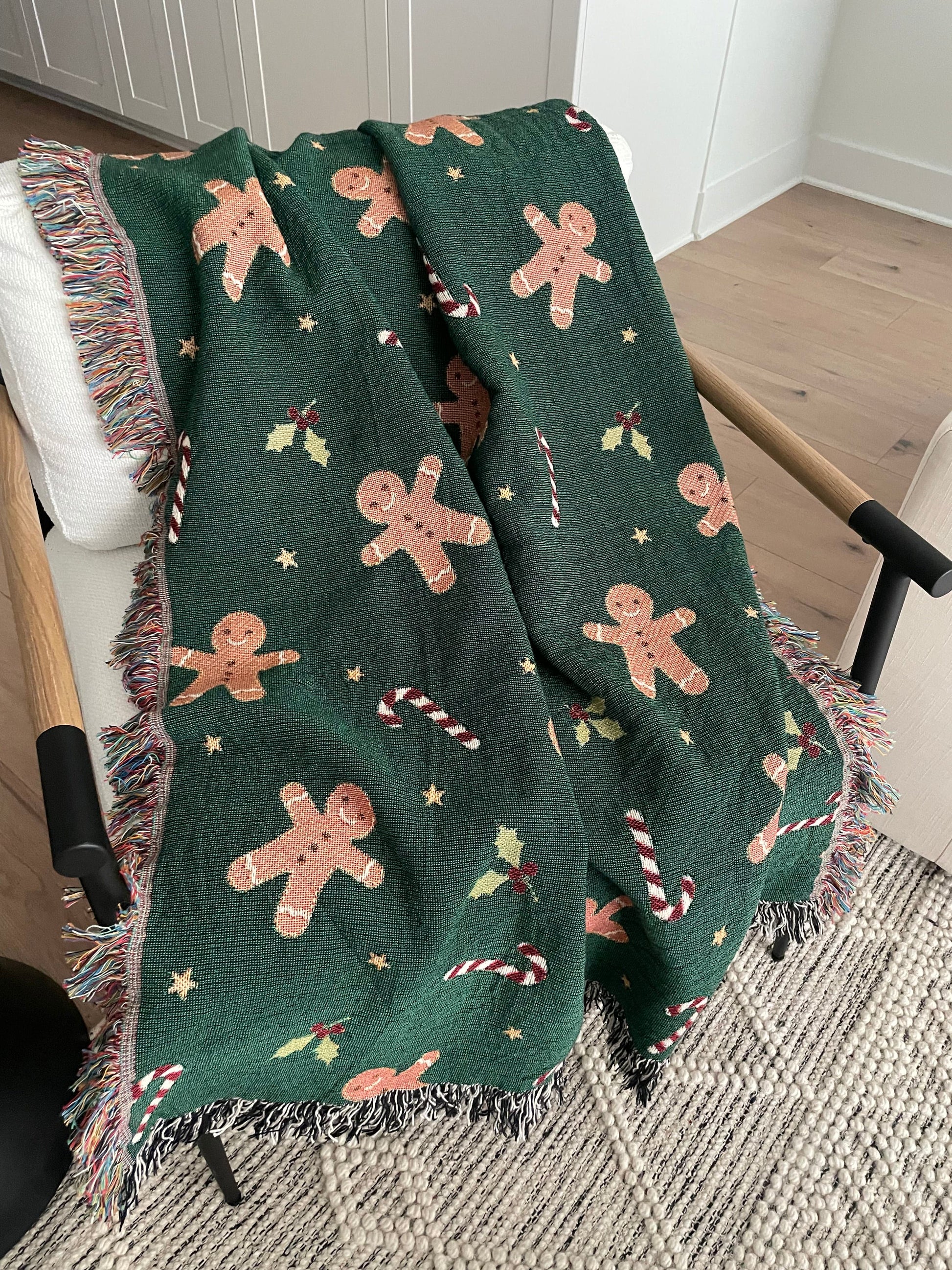 Dark green Christmas woven blanket featuring gingerbread men, candy canes, holly leaves, and multicolored plus black-and-white fringe, draped over a white armchair.